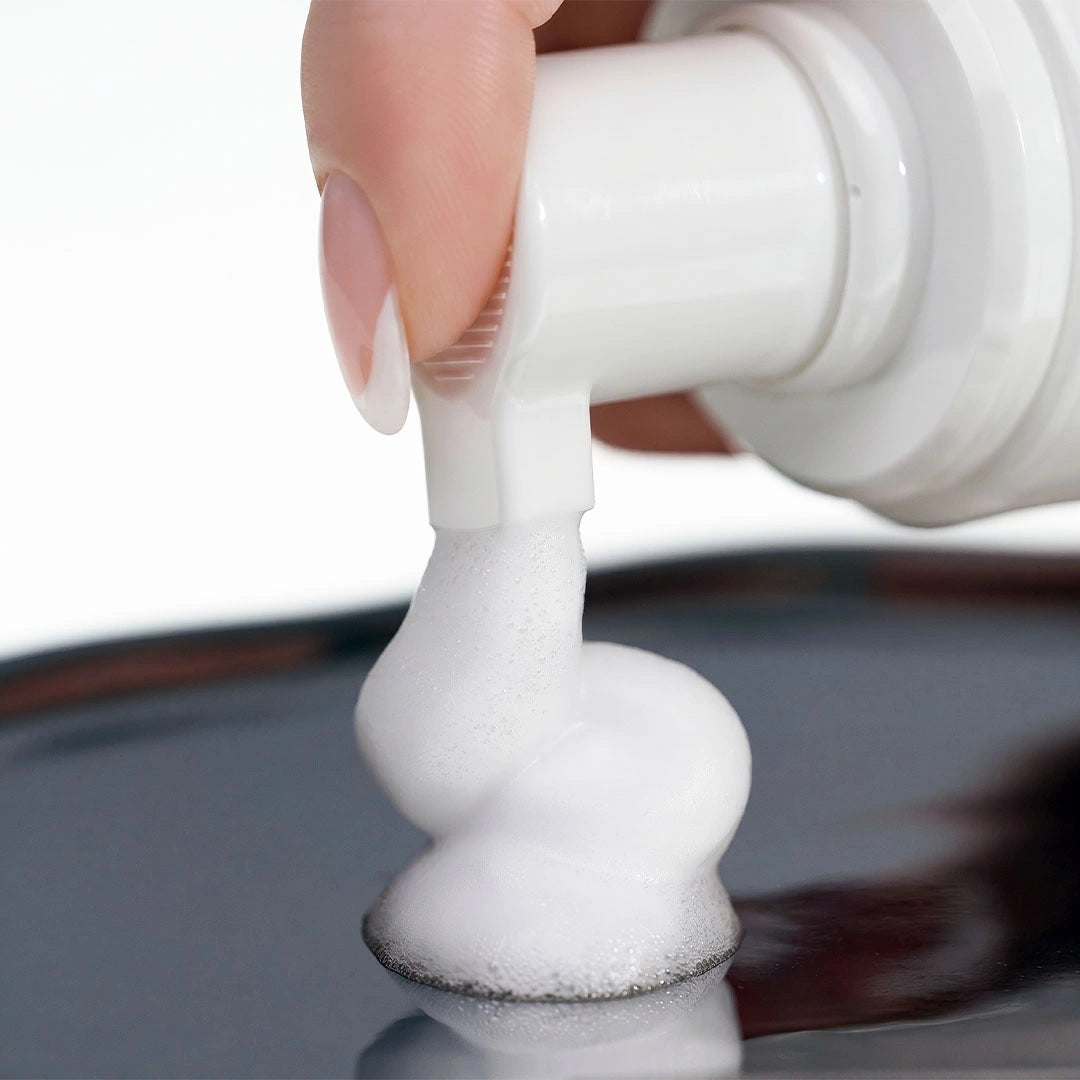 Close-up of the pump dispenser releasing thick white cleansing foam onto a reflective metallic surface.